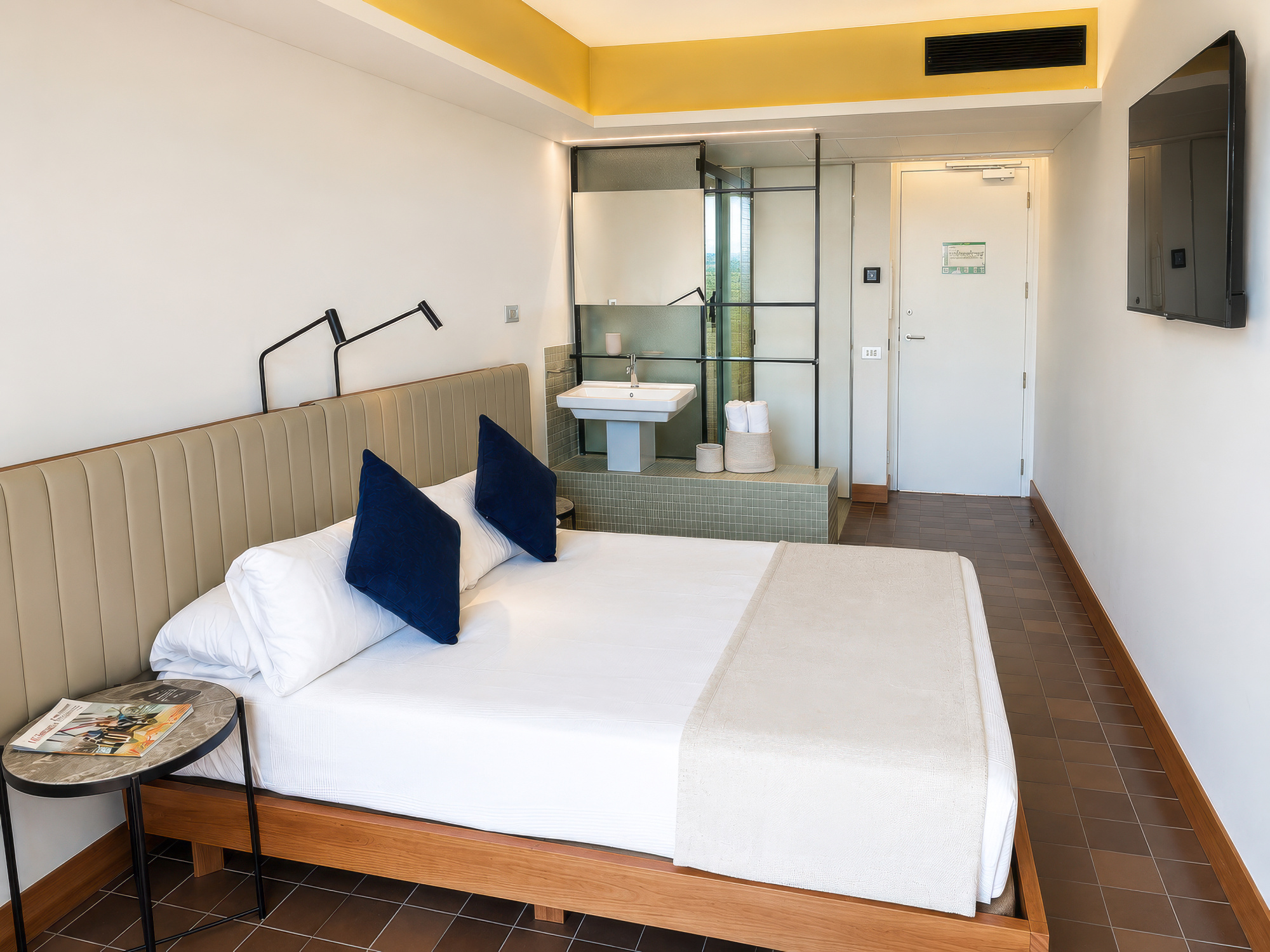 Se ve una cama en una habitación con iluminación empotrada, mesita auxiliar y una zona de cocina al fondo.