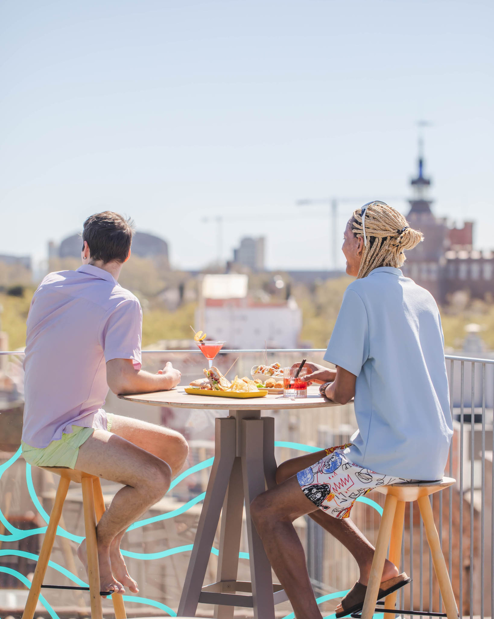 Dos personas se sientan en sillas junto a una mesa con comida en una terraza al aire libre.