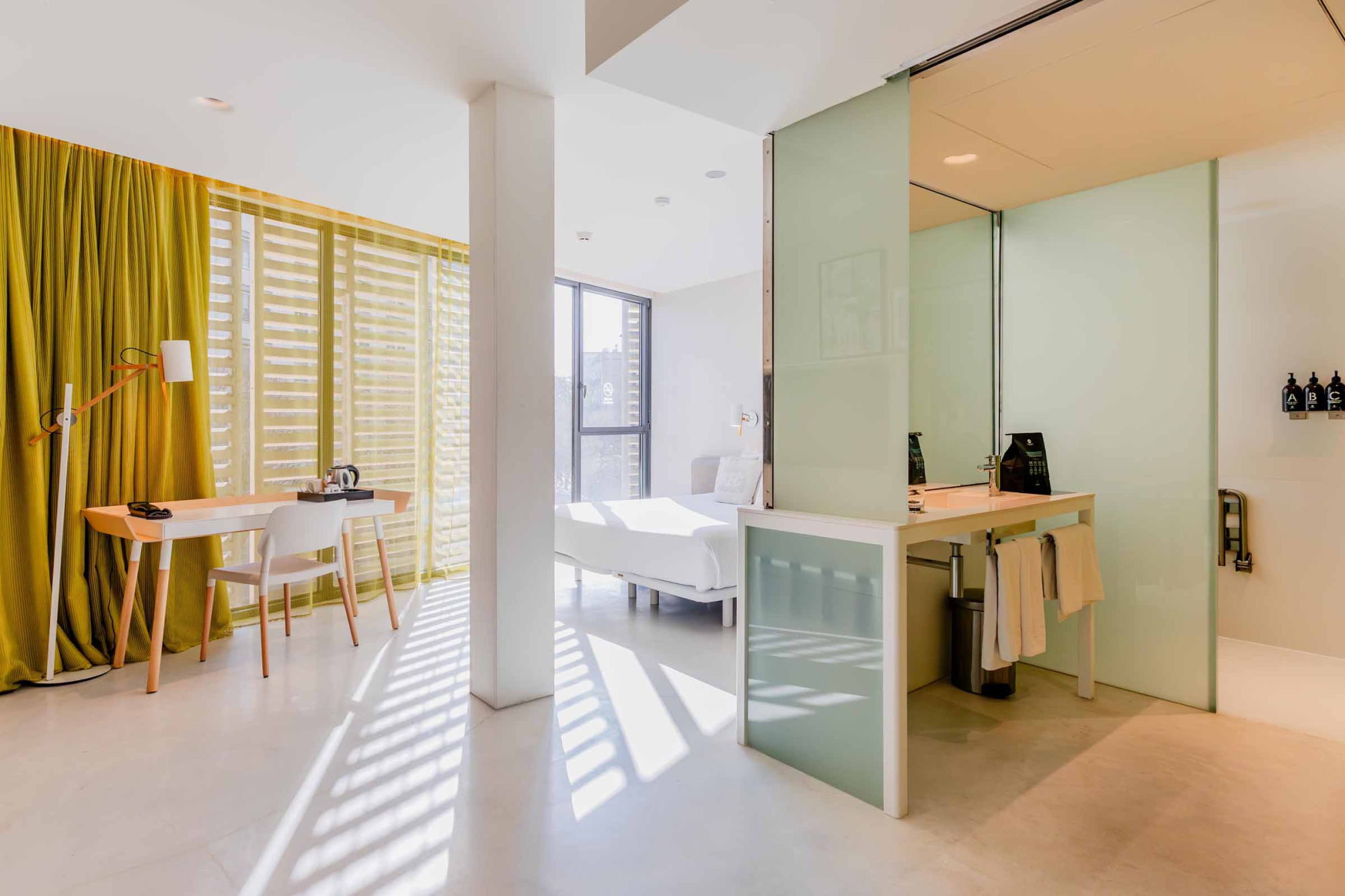 Se ve una sala luminosa con sofá, escritorio y una mesa junto a una ventana con cortinas.