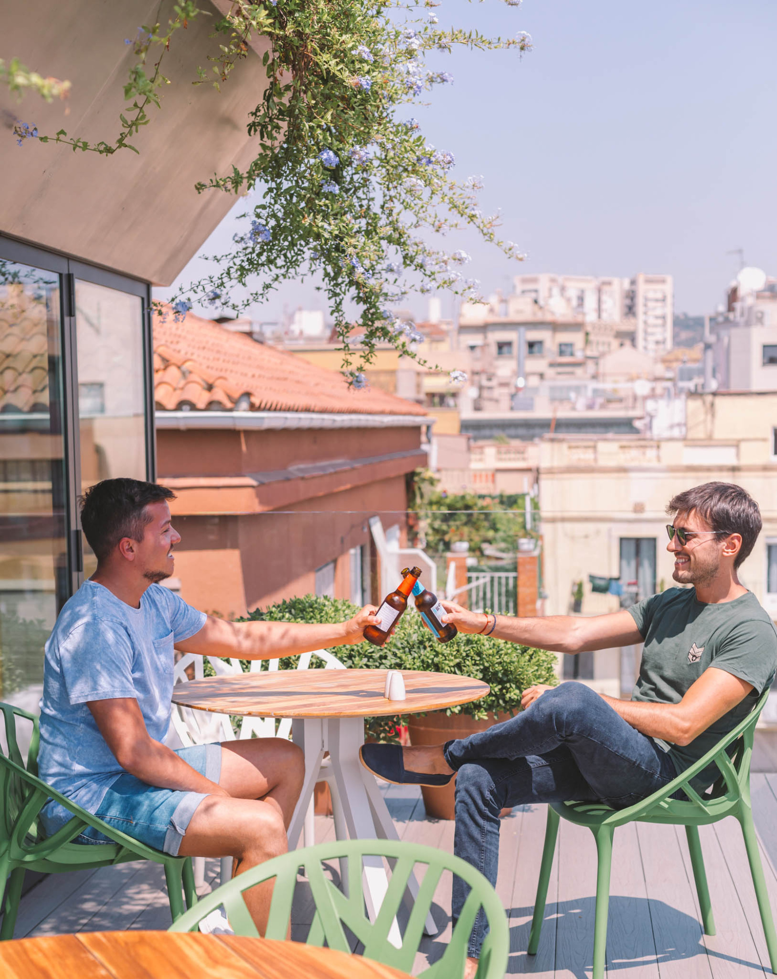Dos personas brindan con botellas sentadas en sillas verdes en una terraza con plantas y vista urbana.