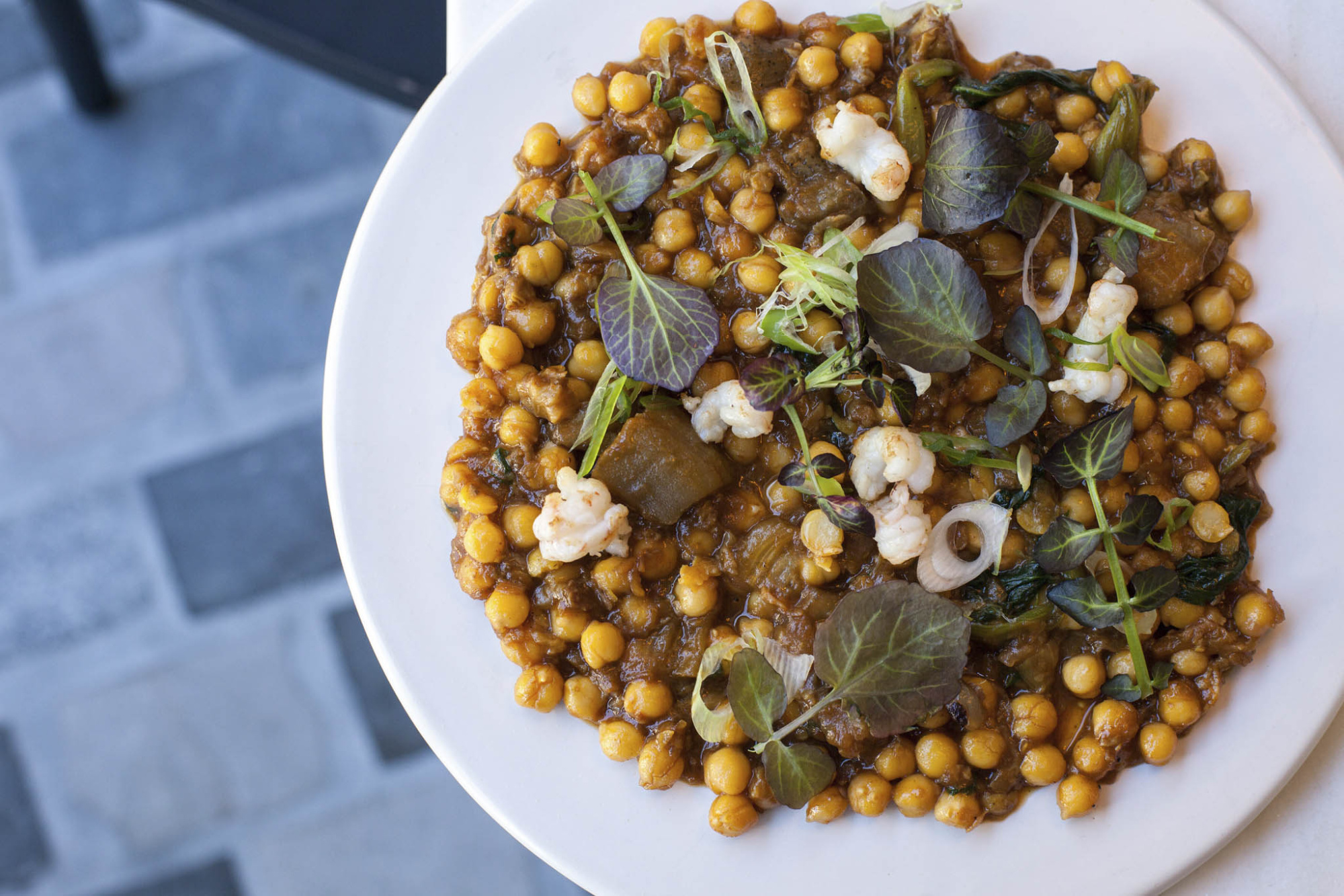 Sirve un plato blanco con garbanzos y hojas verdes sobre una mesa de piedra.