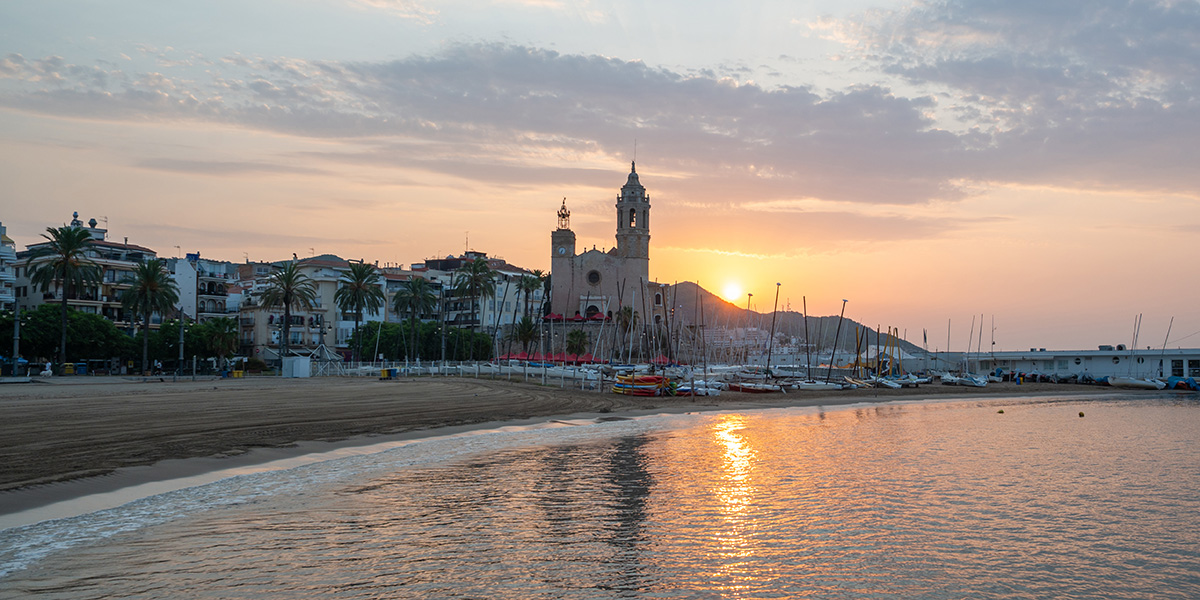 A beach with calm water and moored boats is shown as the sun sets on the horizon.