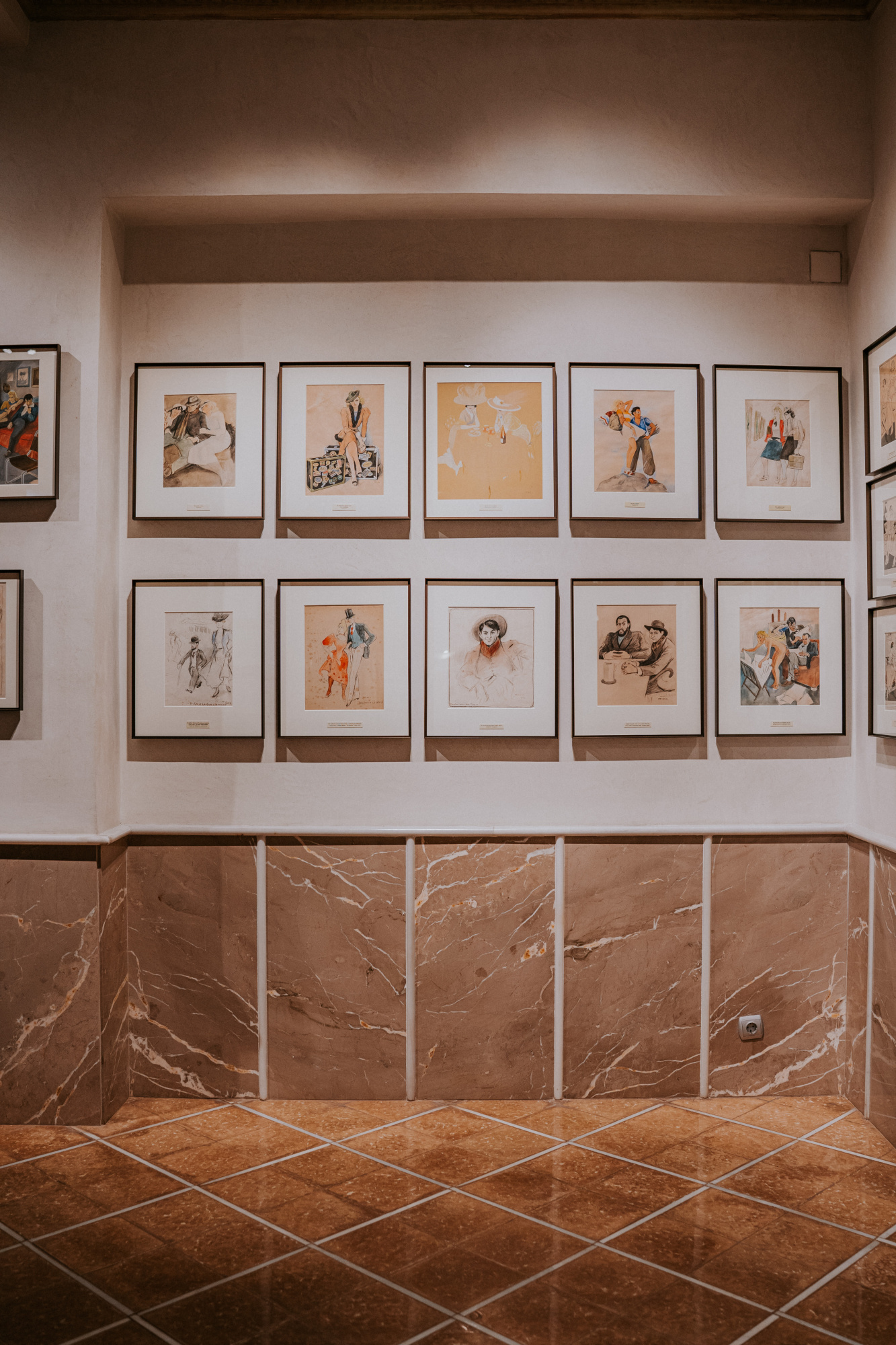 Se ve una pared con varias ilustraciones enmarcadas y un zócalo de azulejos marrones en una sala interior.