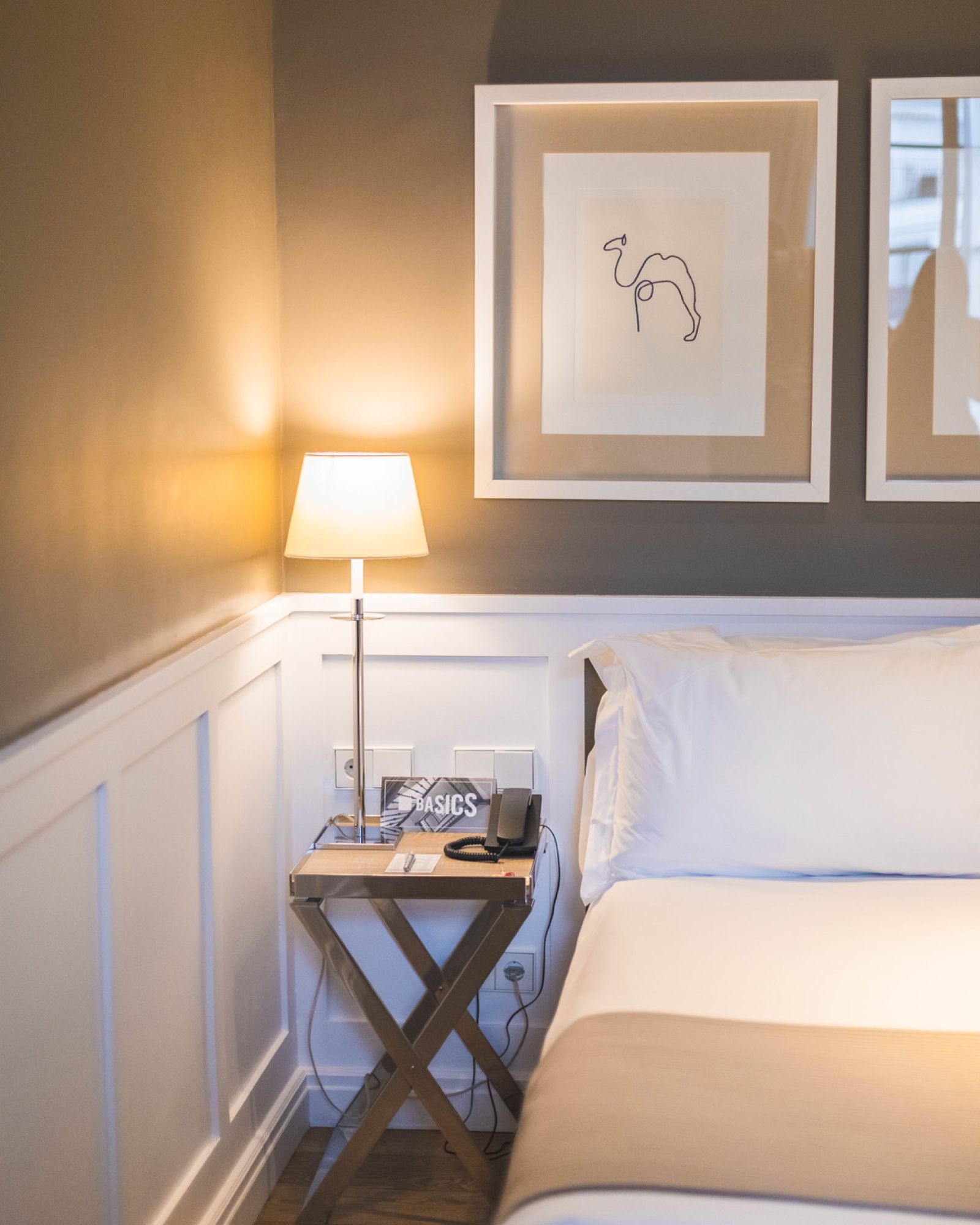 A double bed with light bedding, a side desk, and a lamp can be seen against a lit wall.