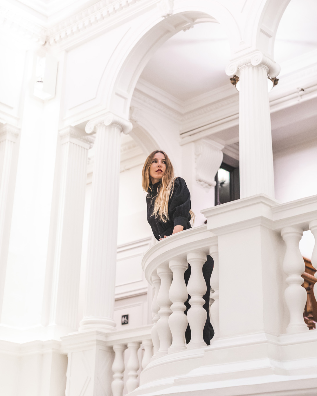 Une femme prend la pose sur un balcon intérieur avec une rambarde blanche et des colonnes ornementales.