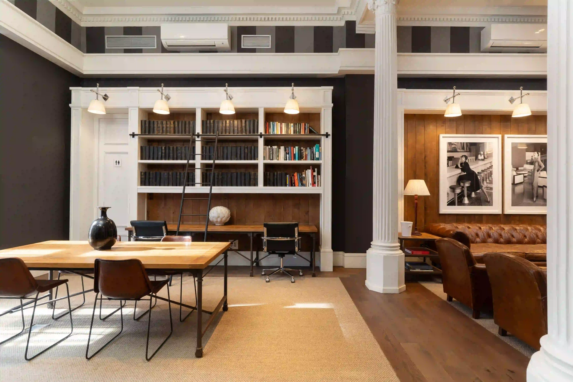 An interior room is shown with bookshelves, work tables and chairs beside white columns.