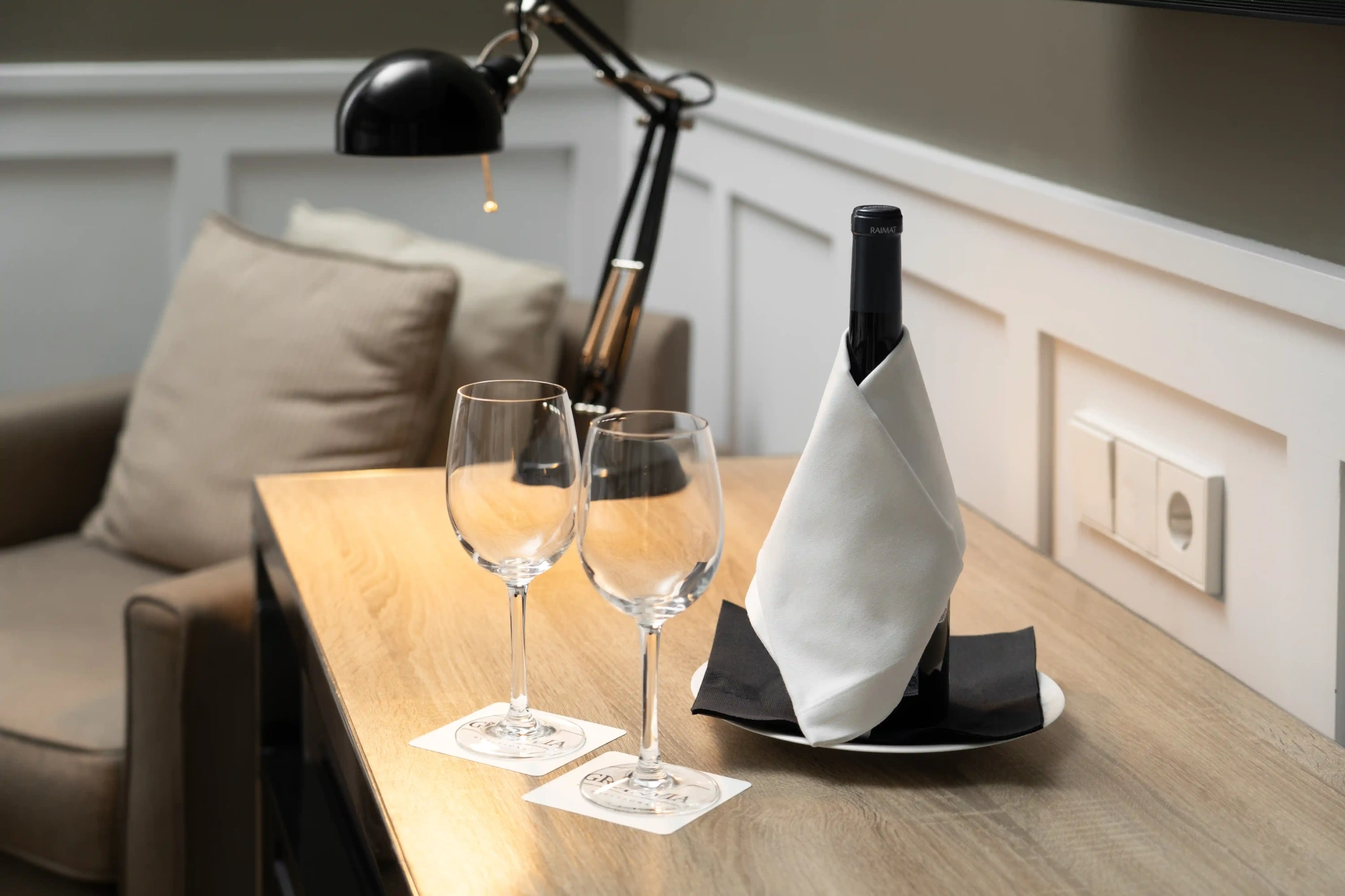 Se ve una mesa de madera con dos copas de vino y una botella sobre una silla junto a un sofá.