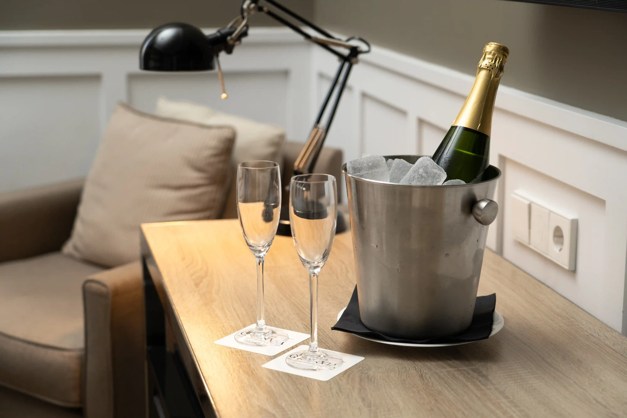 Se ve una mesa con una cubitera metálica con botellas y dos copas sobre un sofá tapizado.