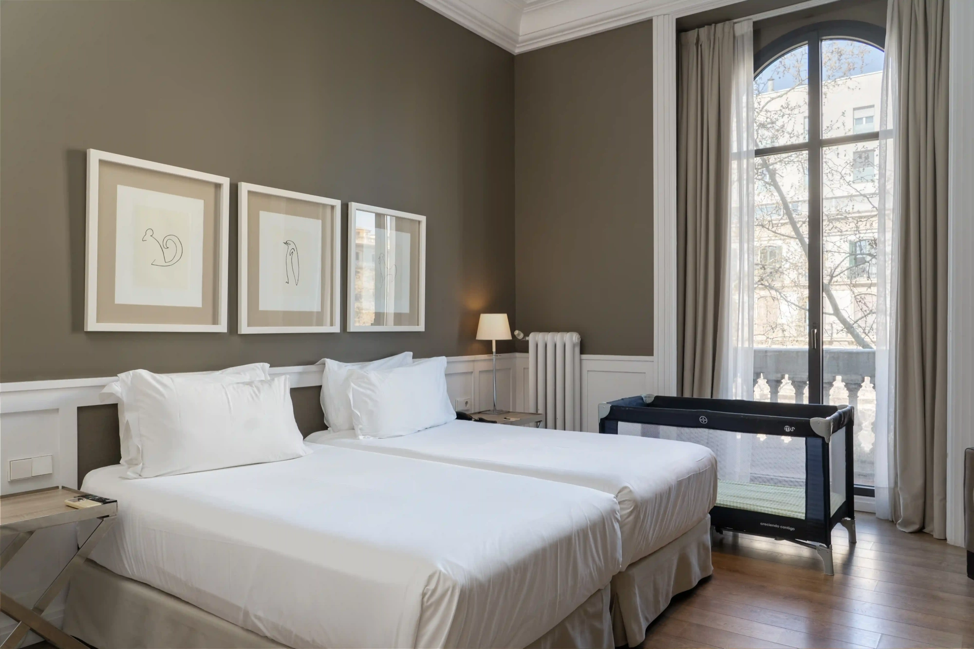 Se ve una cama doble con ropa de cama blanca, mesillas y una ventana con cortinas junto a una zona de estar.