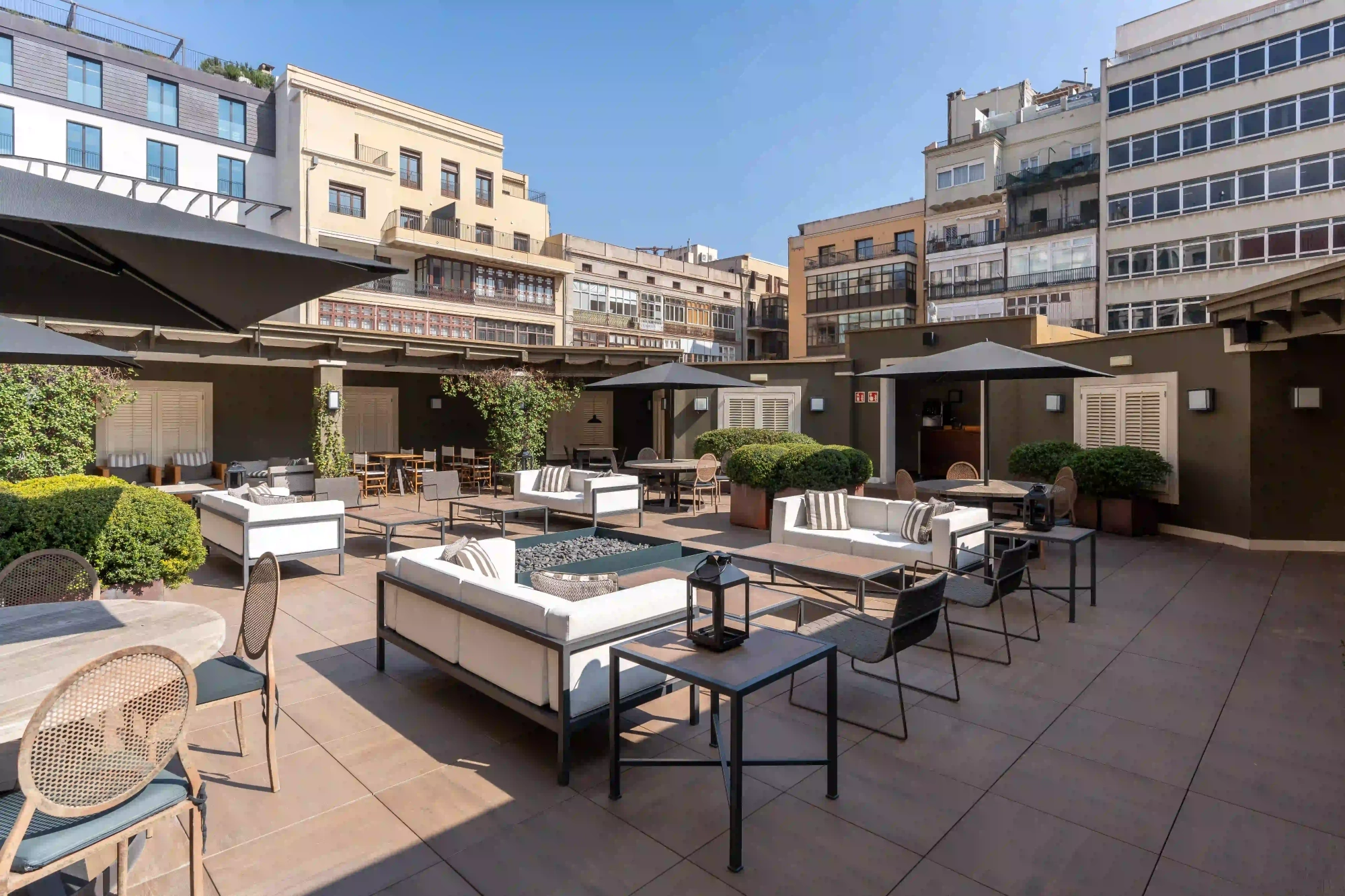 Es veu una terrassa ampla amb taules, cadires i para-sols, envoltada de vegetació i edificis urbans al fons.