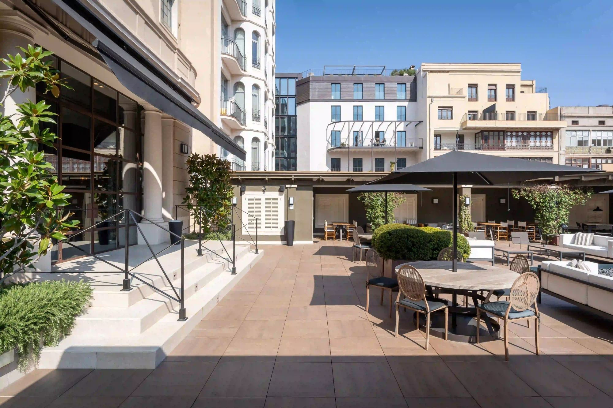 An outdoor terrace is shown with tables and chairs, planters and umbrellas beside a building.
