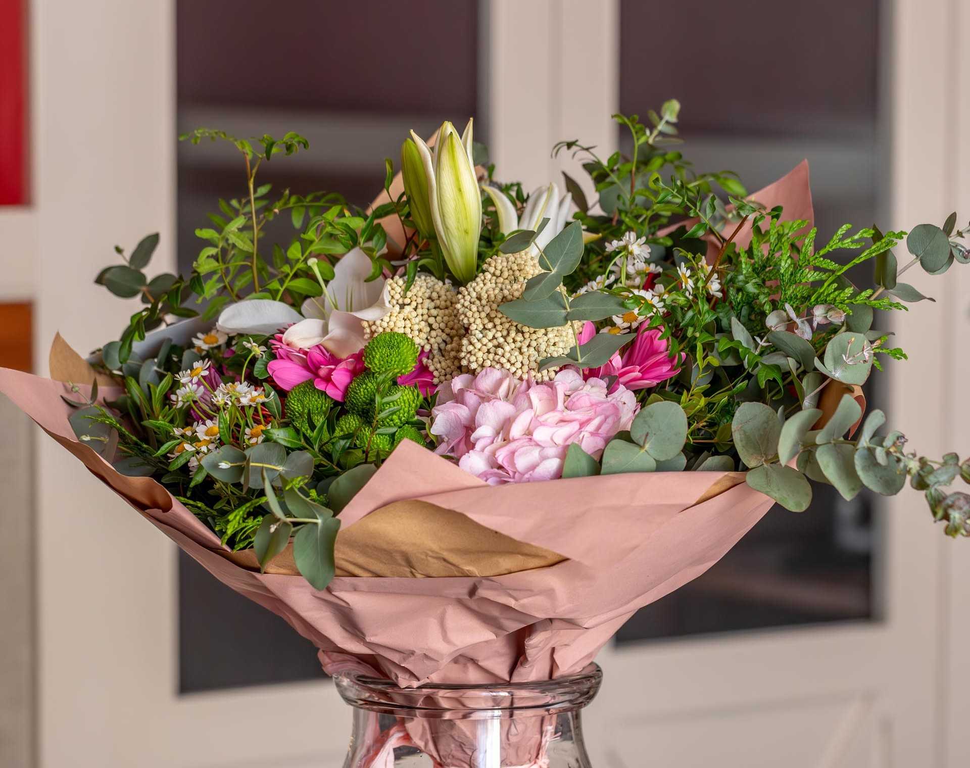 Un ramo de flores envuelto en papel rosa y verde se muestra en un jarrón de vidrio.