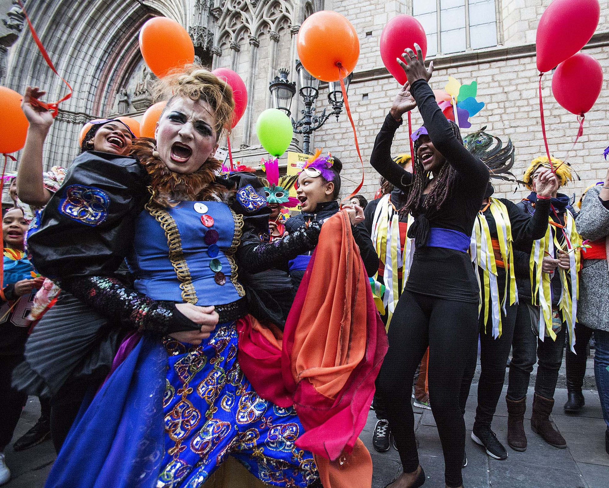 Several people are celebrating in the street with colorful balloons in front of a stone building with arches.
