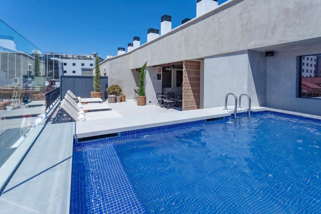 Se ve una piscina azul en una terraza con tumbonas, barandilla y edificios al fondo.