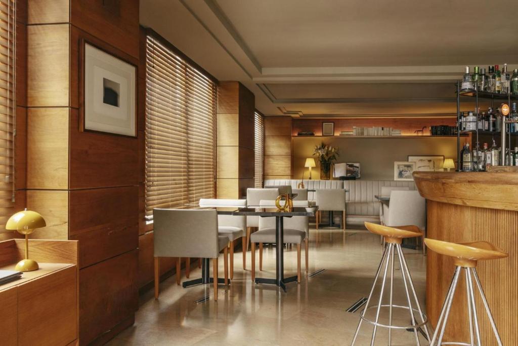 Se ve un salón con barra de madera, taburetes, mesas redondas, asientos tapizados y lámparas de pared.
