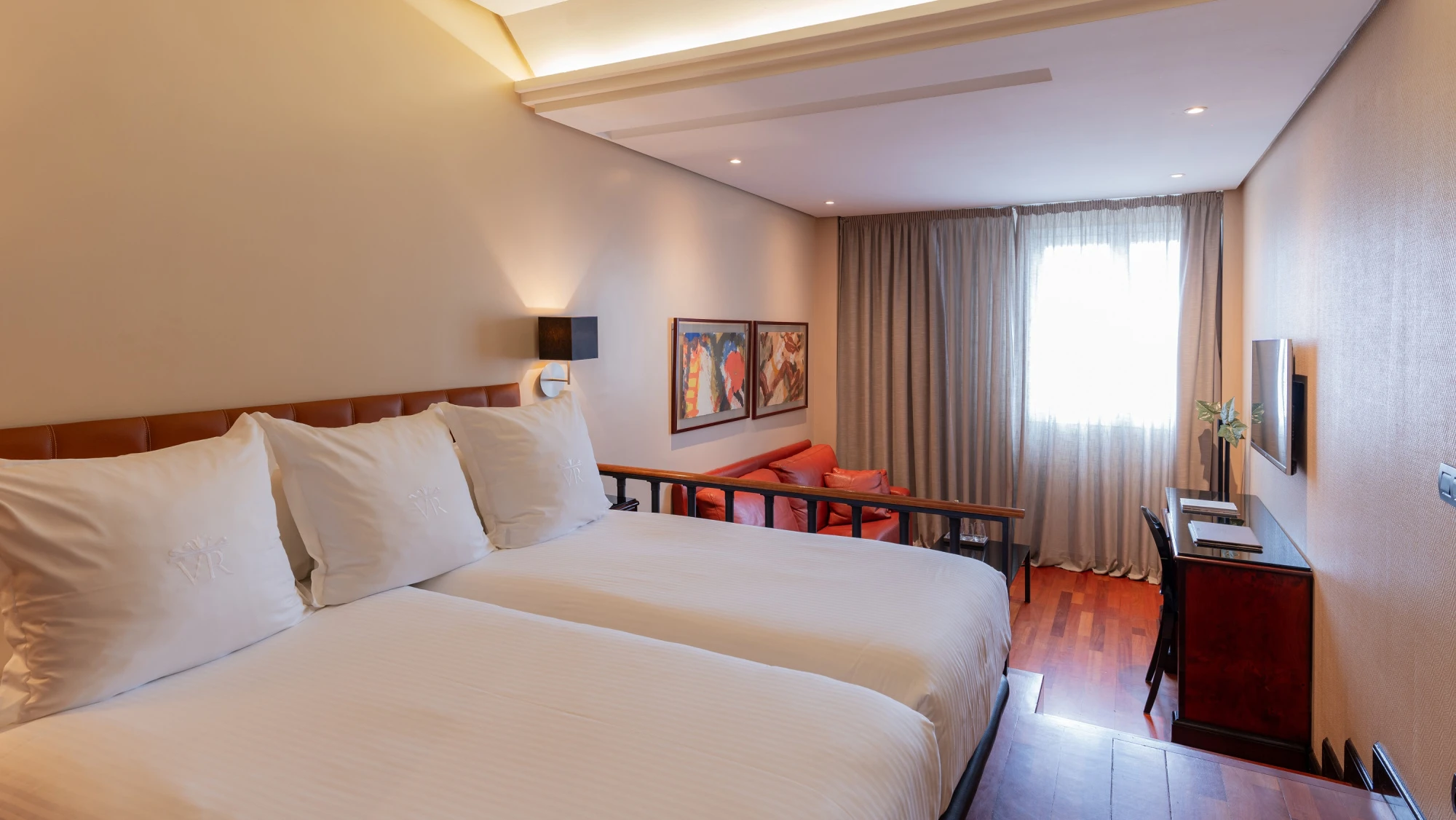 Se ve una cama doble con almohadas, una mesa auxiliar y una ventana con cortinas en una habitación luminosa.
