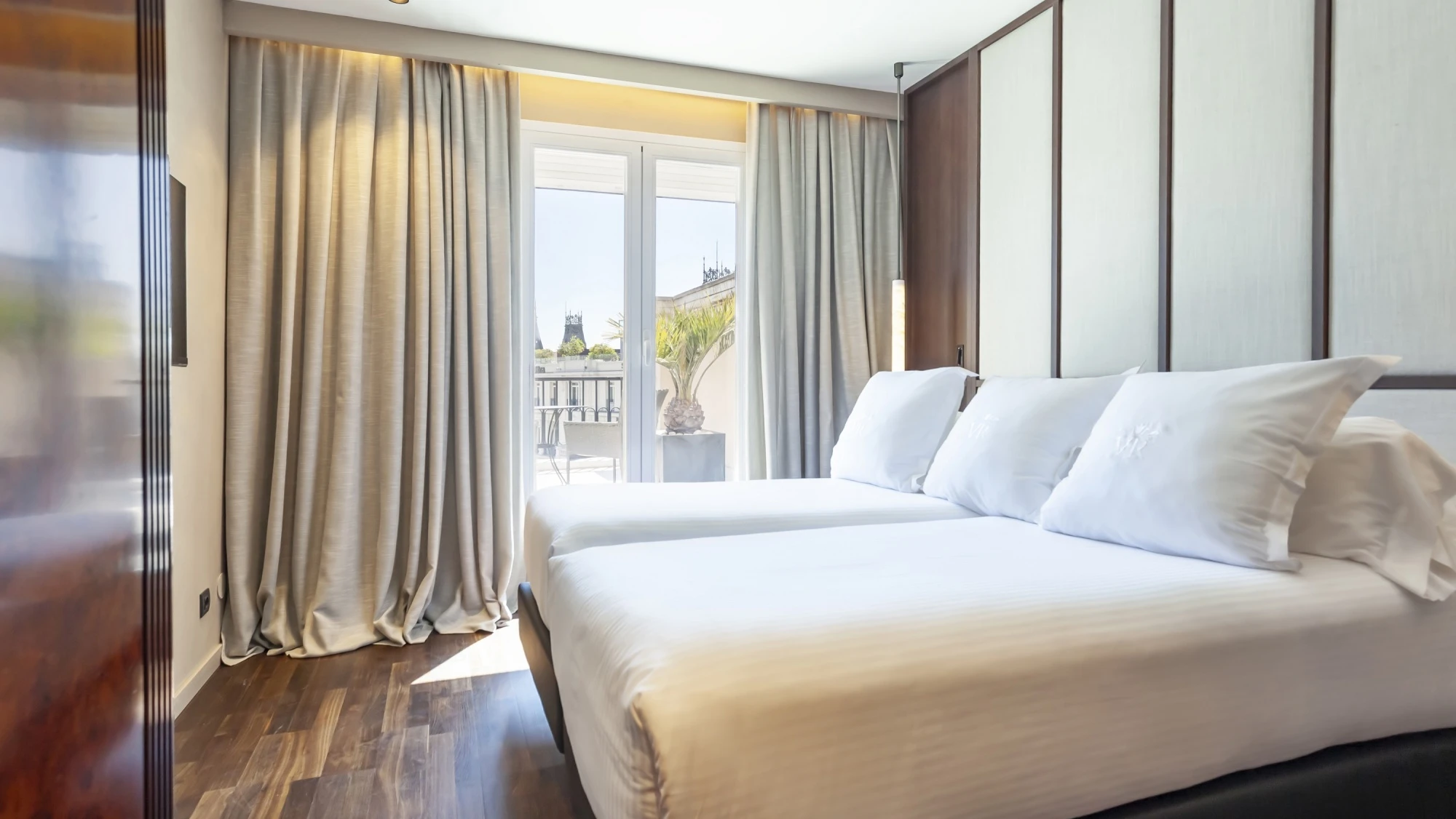 Cama doble con ropa de cama blanca y cortinas junto a un ventanal con salida a un balcón.