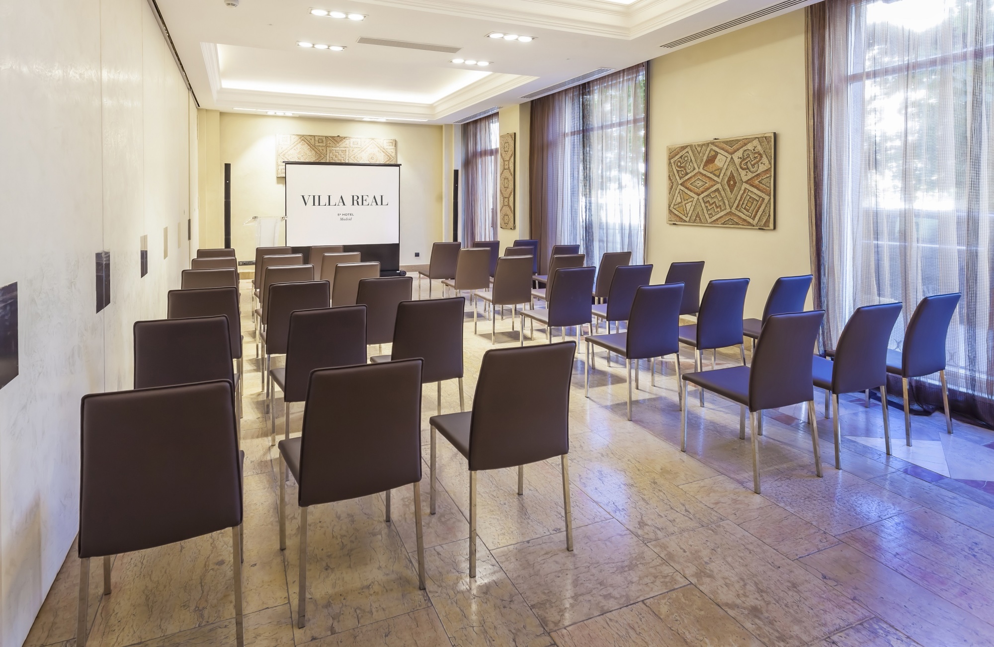 Se ve una sala de reuniones con mesas, sillas y una pared con panel decorativo junto a ventanas con cortinas.