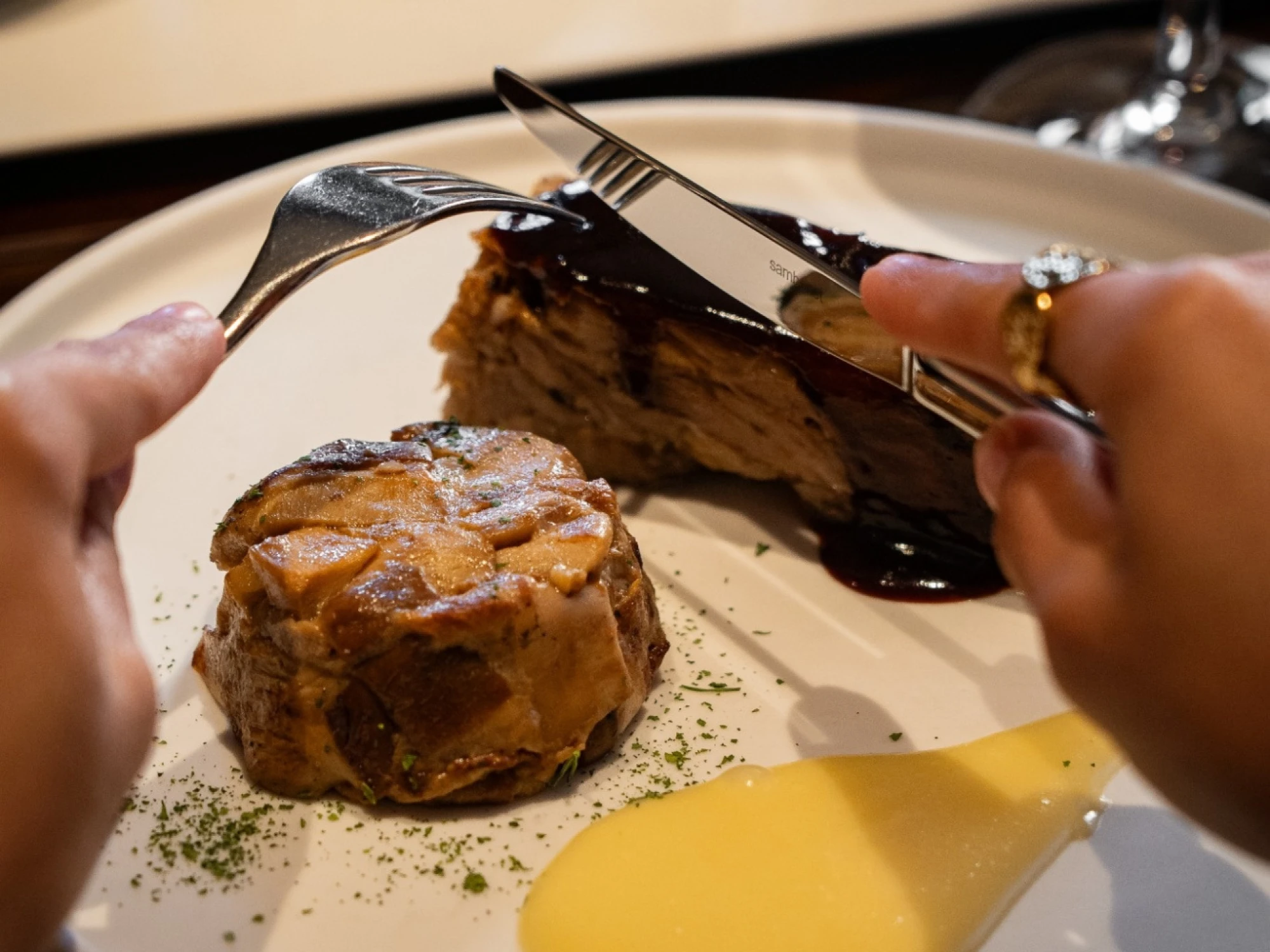 Se ve un plato con comida y cubiertos en una mesa, mientras se corta un bocado con un cuchillo y tenedor.