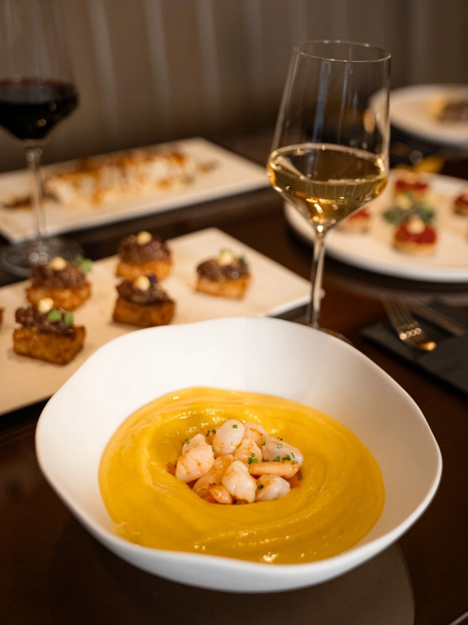 Se ve un plato hondo con crema y marisco, acompañado de una copa de vino sobre una mesa con otros platos al fondo.