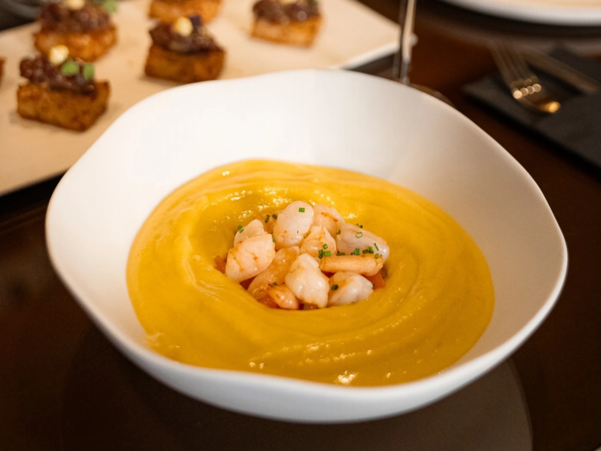 Se ve un plato blanco con crema amarilla y gambas sobre una mesa de restaurante.