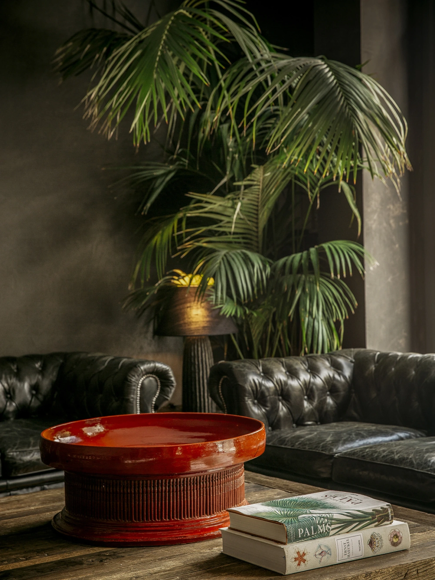 Se ve un sofá tapizado oscuro con una mesa redonda roja y una planta de interior junto a una pared de color oscuro.