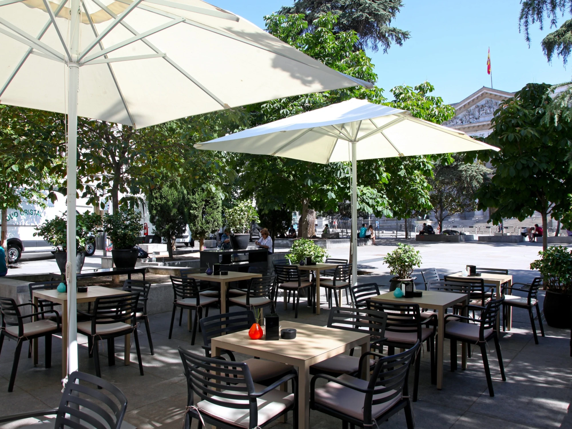 Se ve una terraza exterior con mesas, sillas y sombrillas blancas bajo árboles y cielo despejado.