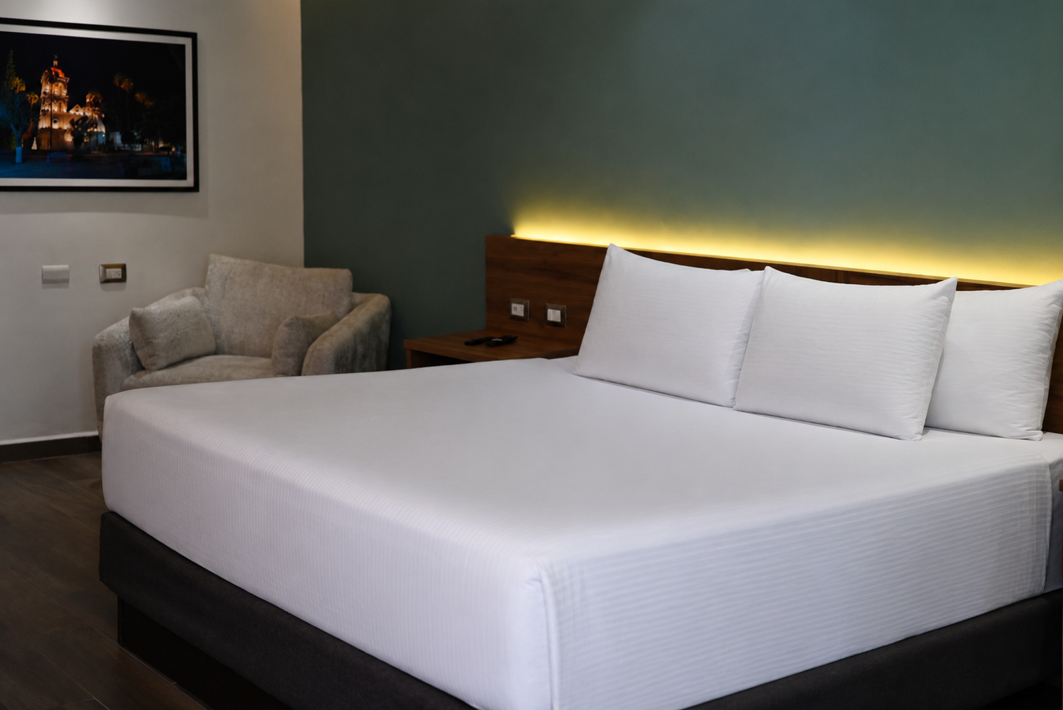 Se ve una cama doble con almohadas blancas y un sofá gris en una habitación con pared verde.