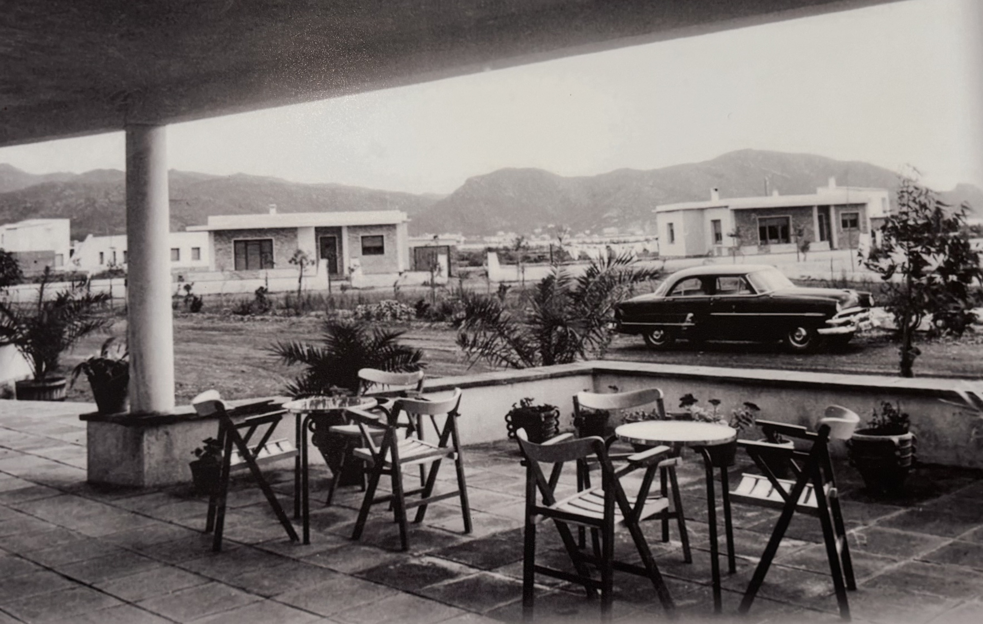 Se ve una terraza con mesas y sillas, y un coche estacionado frente a edificios.