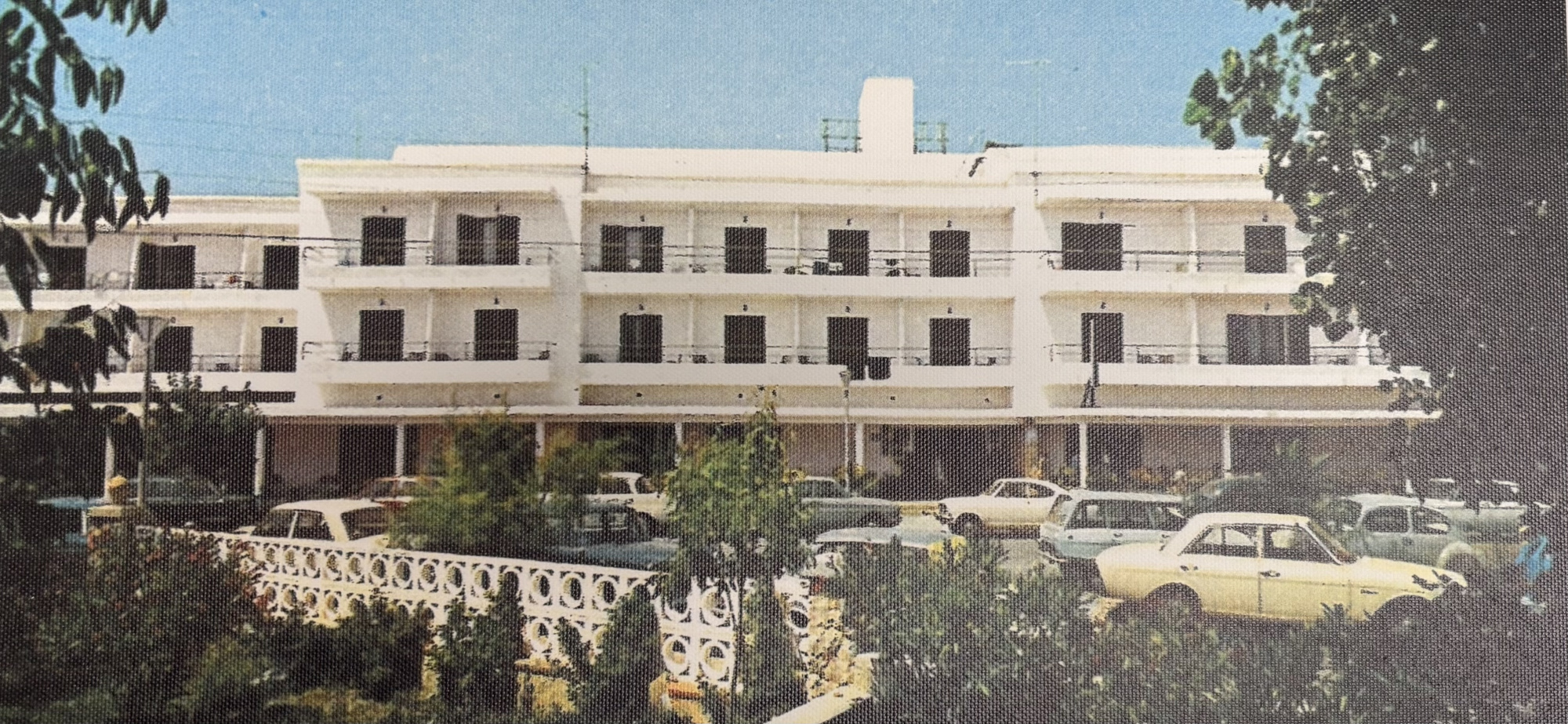 Se ve la fachada de un edificio blanco con balcones y varios coches estacionados frente a él.
